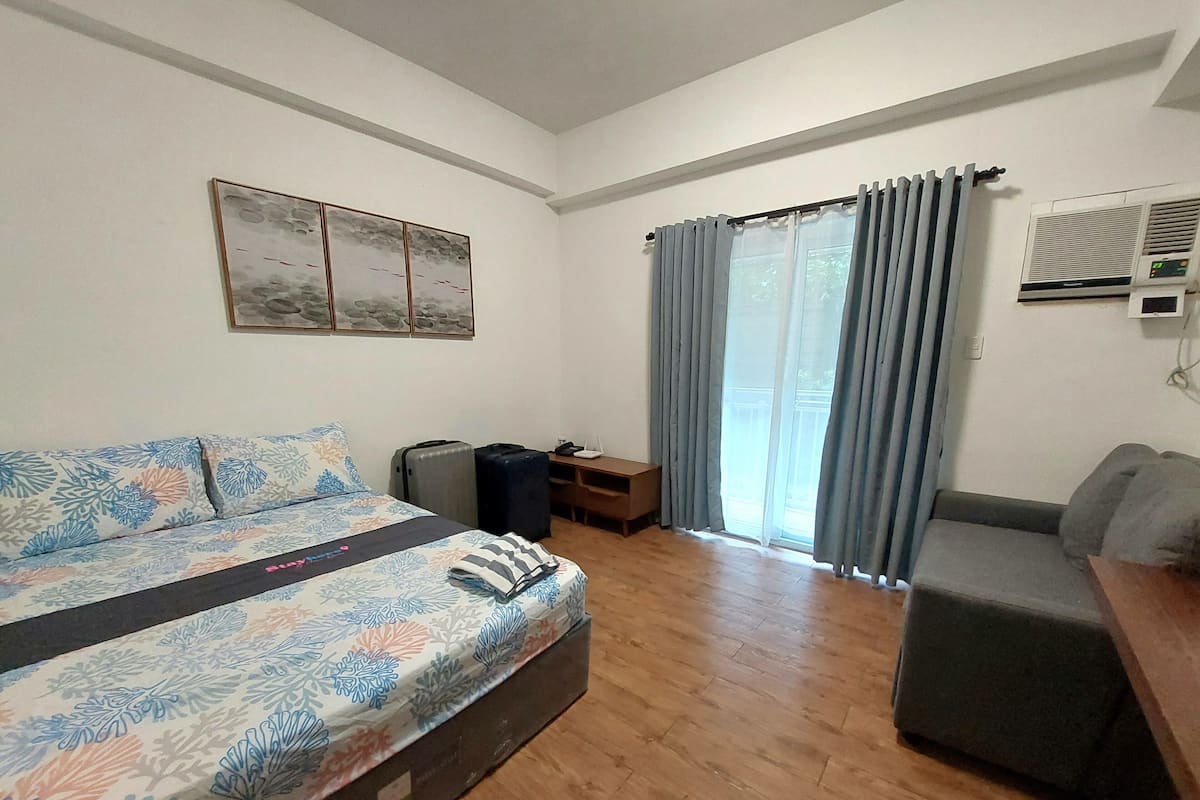 Quarto de hotel com cama, sofá, escrivaninha, mala e janela com cortinas.