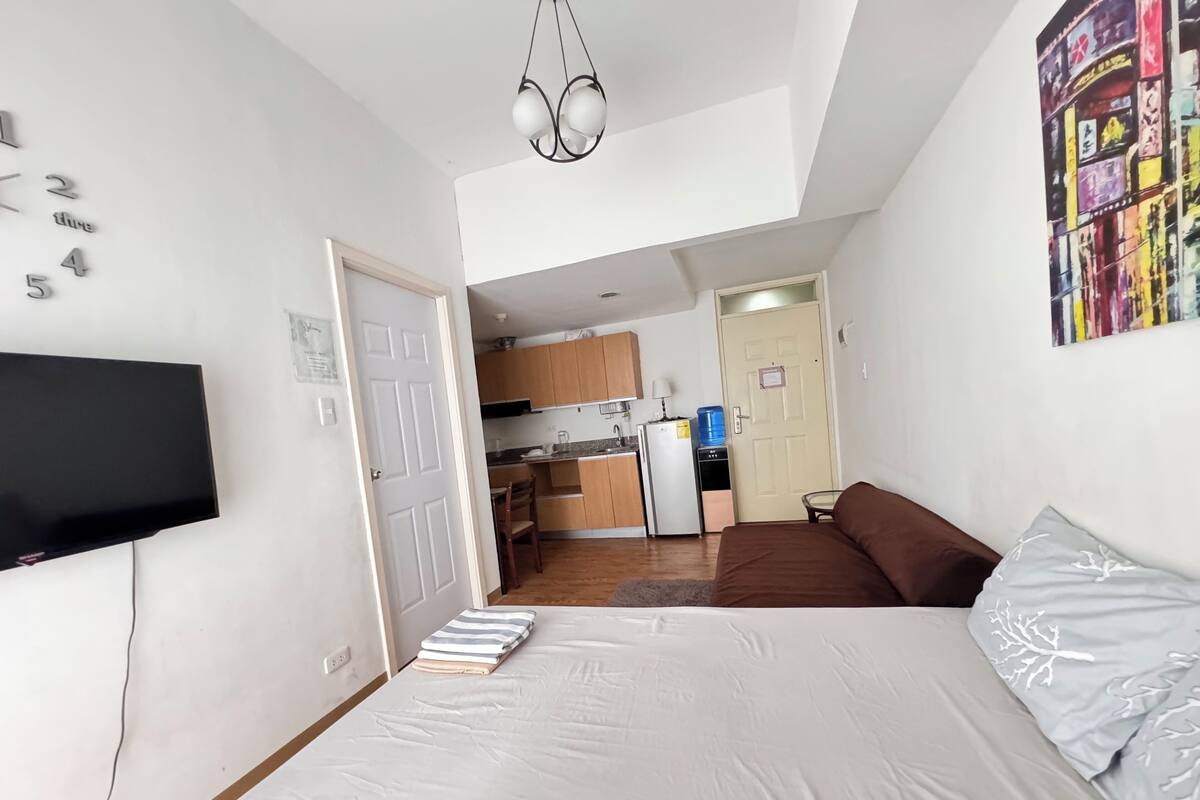 Quarto com uma cama, televisão na parede, cozinha compacta ao fundo e uma porta que dá para outro espaço.