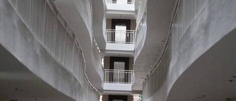 Corredor de hotel em vários níveis, com varandas, plantas em vasos e uma placa de saída.