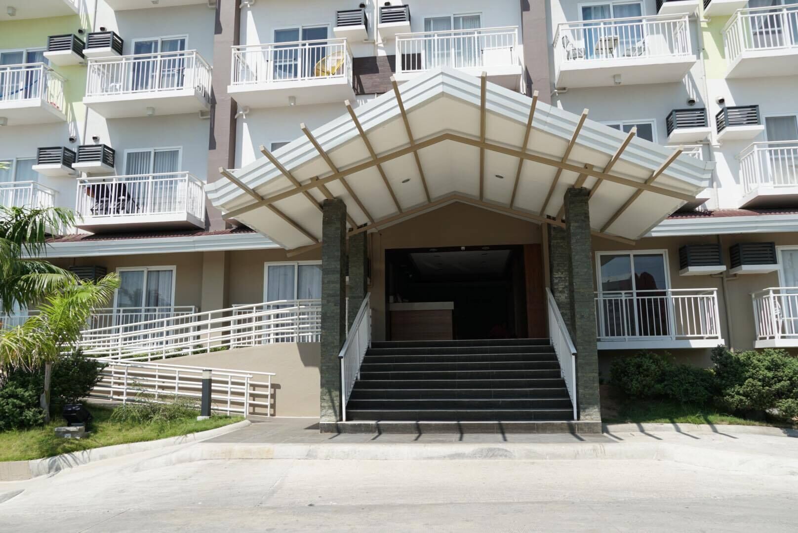 A multi-story hotel entrance with a covered staircase, outdoor seating, and a palm tree.