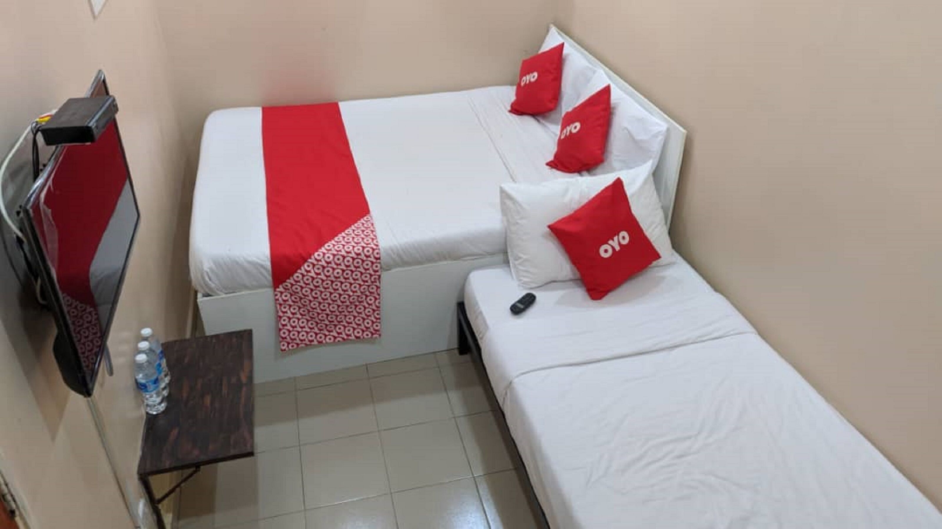 Une petite chambre d’hôtel avec deux lits, chacun agrémenté d’un oreiller rouge et d’une couverture. Il y a une télévision sur le côté gauche et une petite table avec une bouteille d’eau.