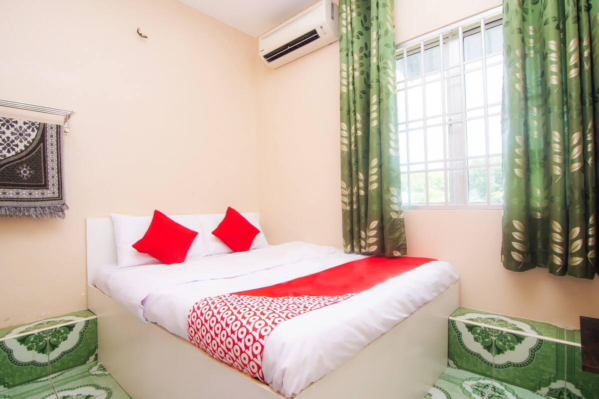 A neatly made bed with red and white bedding, a patterned wall hanging, and a window with green curtains.