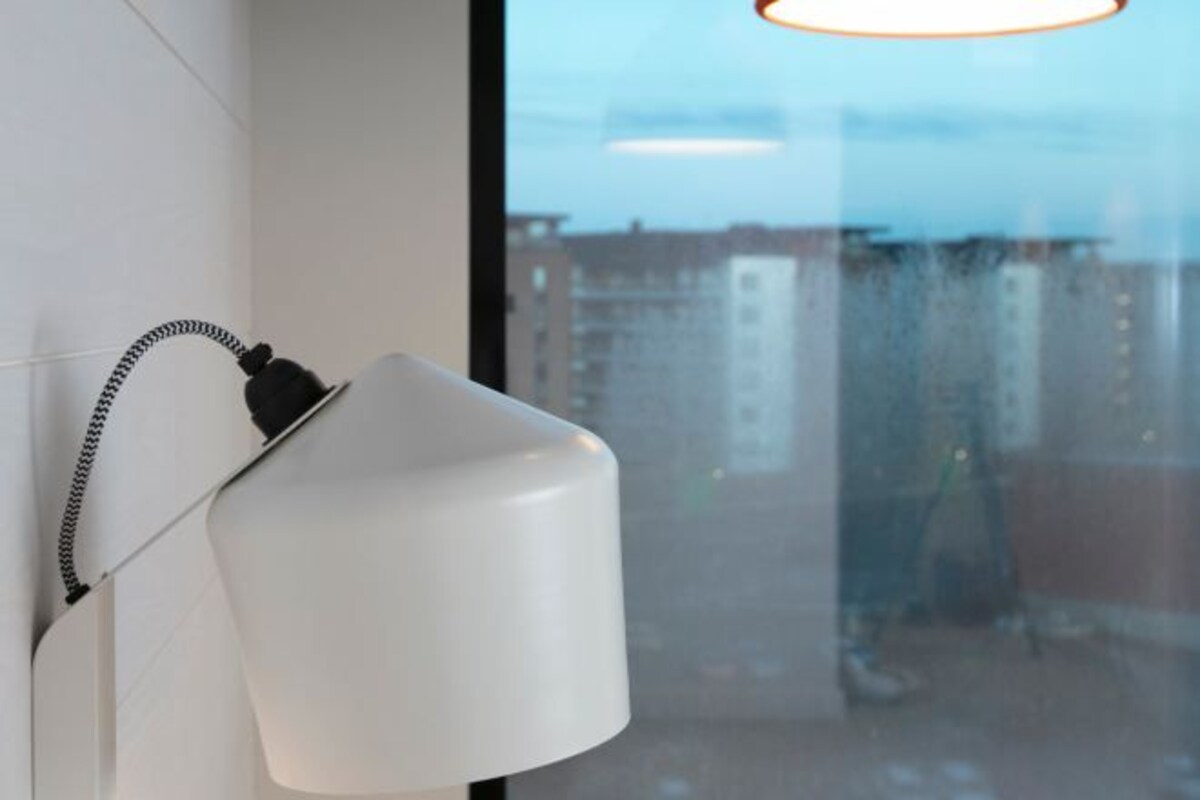 A white wall-mounted lamp with a coiled cord, a window with a view of buildings, and a glass surface reflecting the interior.