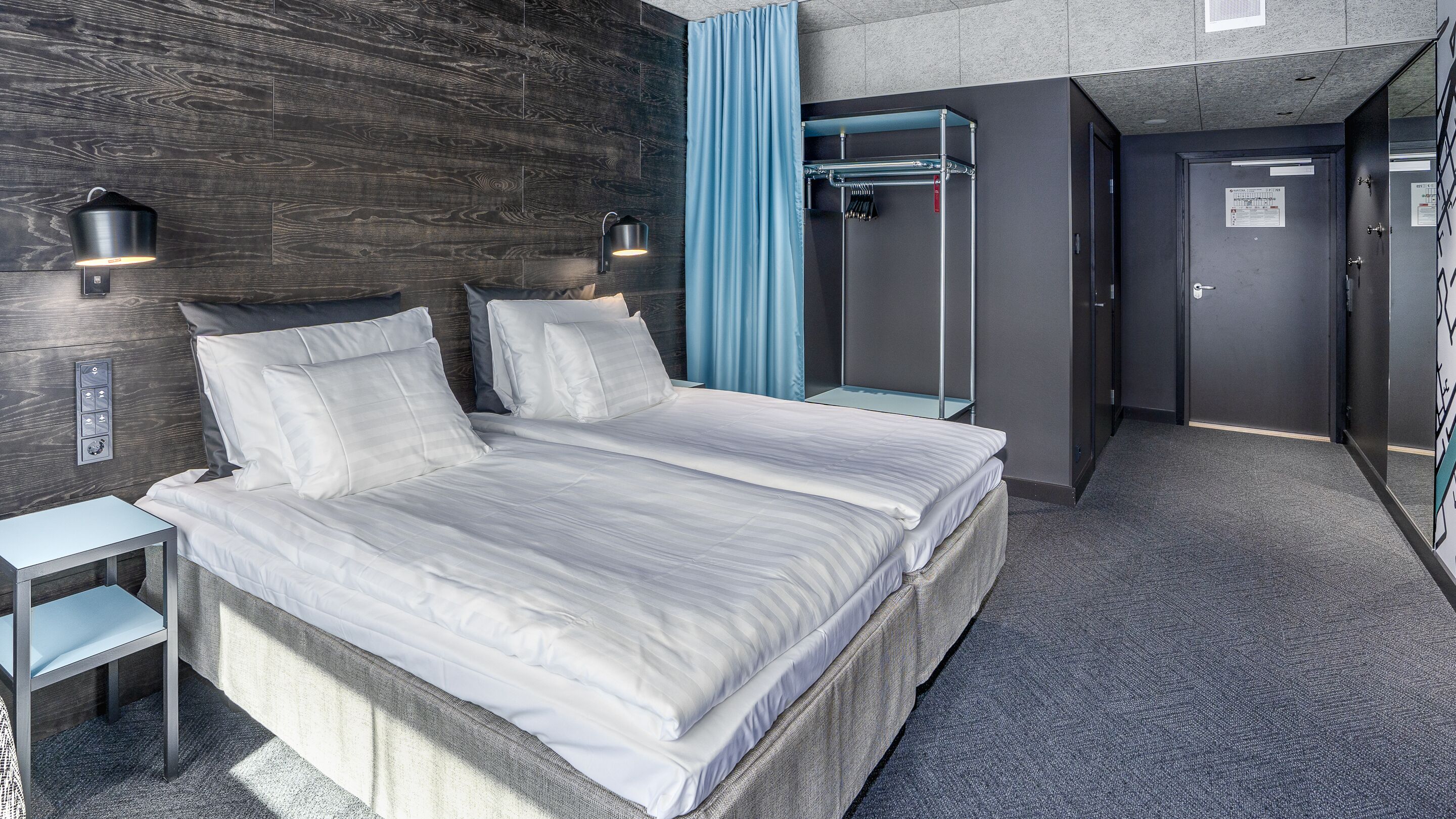 Moderni hotellihuone, jossa on suuri sänky, kaksi yöpöytää, vaatekaappi sekä kylpyhuone, jossa on suihku ja pyyhekoukku.