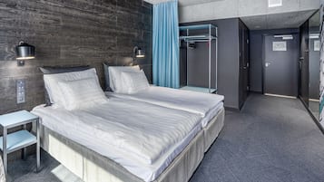 Moderni hotellihuone, jossa on suuri sänky, kaksi yöpöytää, vaatekaappi sekä kylpyhuone, jossa on suihku ja pyyhekoukku.