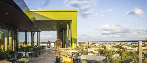 Una terraza al aire libre con mesas y sillas, vistas a la ciudad y un edificio moderno con fachada verde.