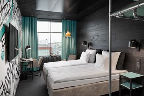 A modern hotel room with a large bed, a desk, and a chair. There is a window with curtains, a wall-mounted TV, and a lamp on a bedside table. - Original Sokos Hotel Kupittaa (Turku)