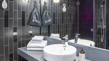 A modern bathroom with a large mirror, a round sink, and a shower. There are towels and a trash can on the counter.