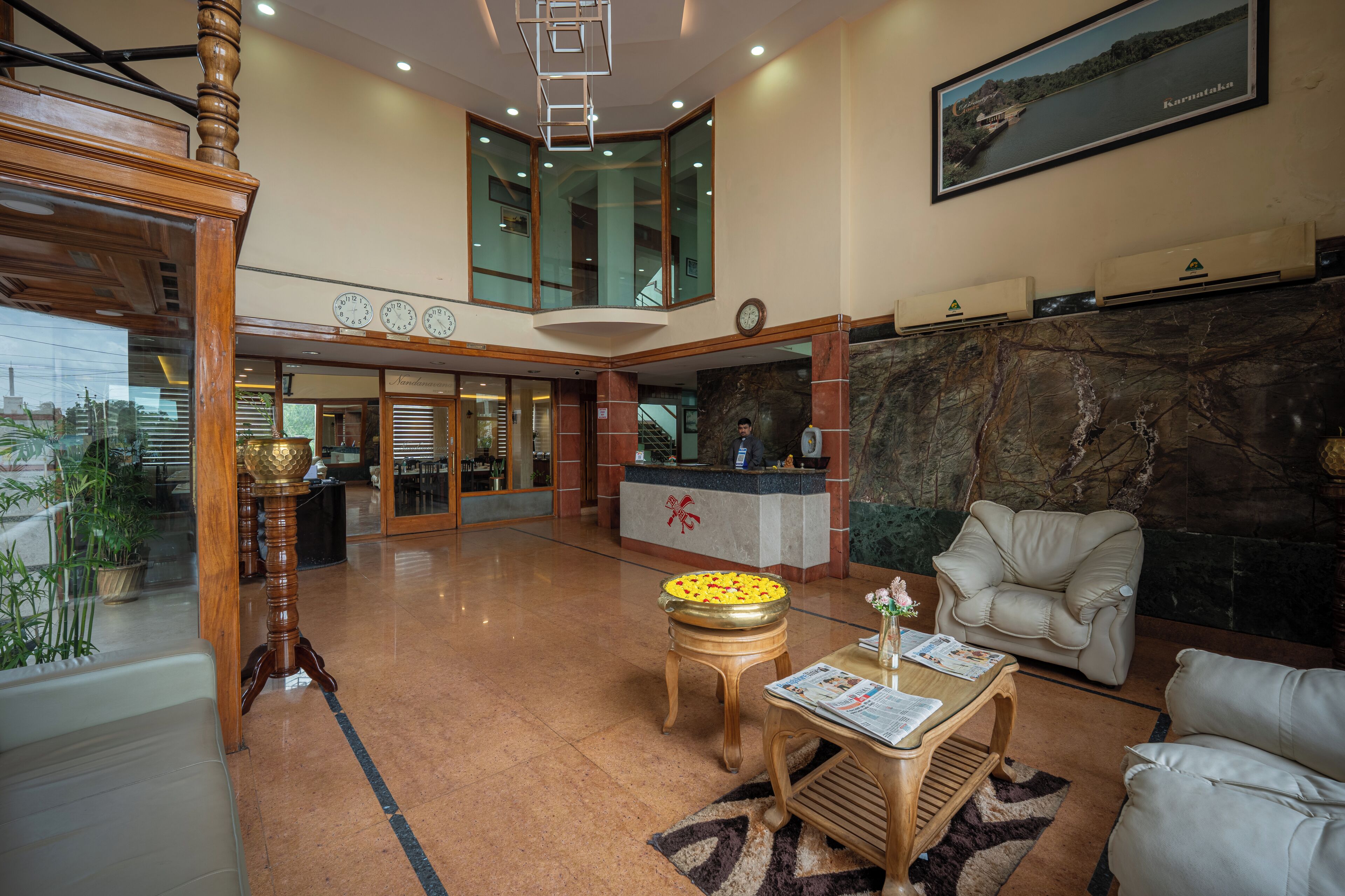 A hotel lobby with a reception desk, a seating area with a sofa and armchairs, a decorative table with a flower vase, and a large window with a view of a body of water.