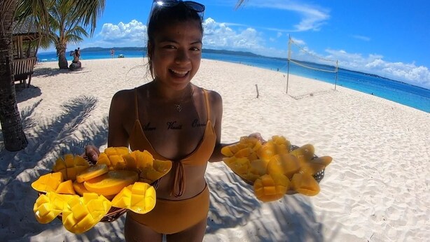 A person holding two baskets of mangoes on a beach with palm trees and a clear blue sky in the background. - Secret Spot Siargao - Hostel (General Luna)