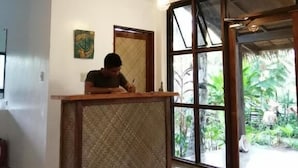 A room with a reception desk, a ceiling fan, a large window with a view of greenery, and a wall-mounted light fixture. - Secret Spot Siargao - Hostel (General Luna)