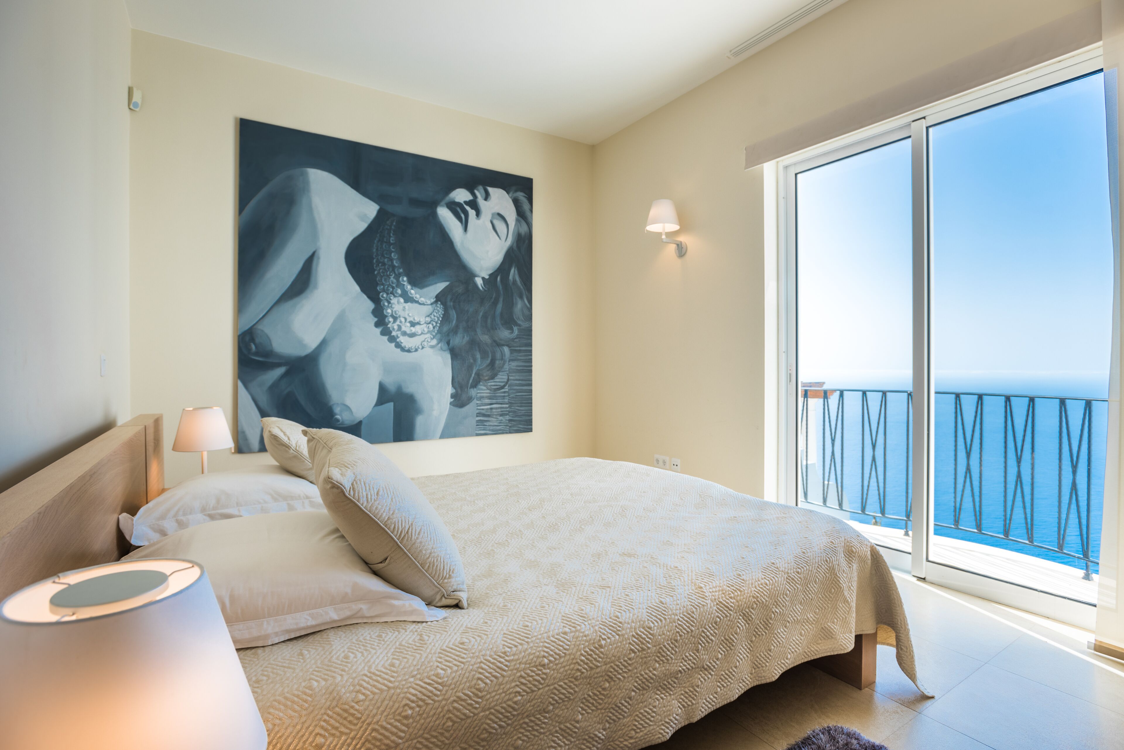 A bedroom with a large bed, a painting on the wall, a nightstand, and a view of the sea through a balcony.
