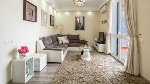 Un salon moderne avec un canapé d’angle, une table basse en verre et un tapis moelleux. Des tableaux encadrés ornent le mur, ainsi qu’une lampe et une fenêtre avec des rideaux.