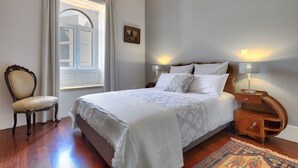 Un lit bien fait, recouvert d’une courtepointe blanche, une table de chevet en bois avec une lampe, une chaise et une fenêtre avec des rideaux.