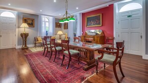 Un coin repas avec une longue table en bois, des chaises à revêtement vert et un tapis rouge à motifs. On y trouve deux lampes, un tableau accroché au mur et une porte avec un panneau indiquant la sortie.