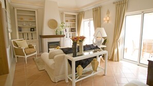 A living room with a fireplace, a sofa, a coffee table, and a dining area with chairs. - Dream Luxury Apartment  Palheiro Village (Funchal)