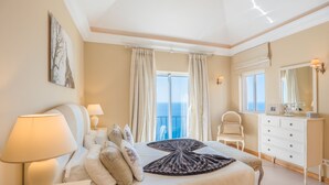 A bedroom with a bed, bedside lamps, a dresser, a mirror, and a view of the sea through the window. - Dream Luxury Apartment  Palheiro Village (Funchal)