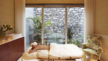 Relax in this serene spa room with a view of lush greenery, featuring a massage table and a comfortable chair.