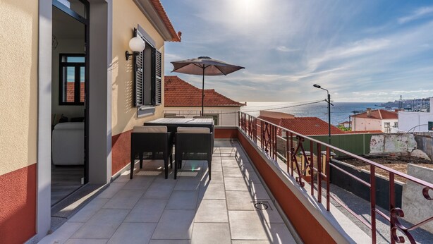 A balcony with a table and chairs, a view of the sea, and a building with a red roof. - Lazareto Apartment (Funchal)