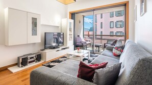 Ein modernes Wohnzimmer mit Fernseher, Couchtisch und grauem Sofa mit roten Kissen. Ein großes Fenster bietet Blick auf Gebäude.