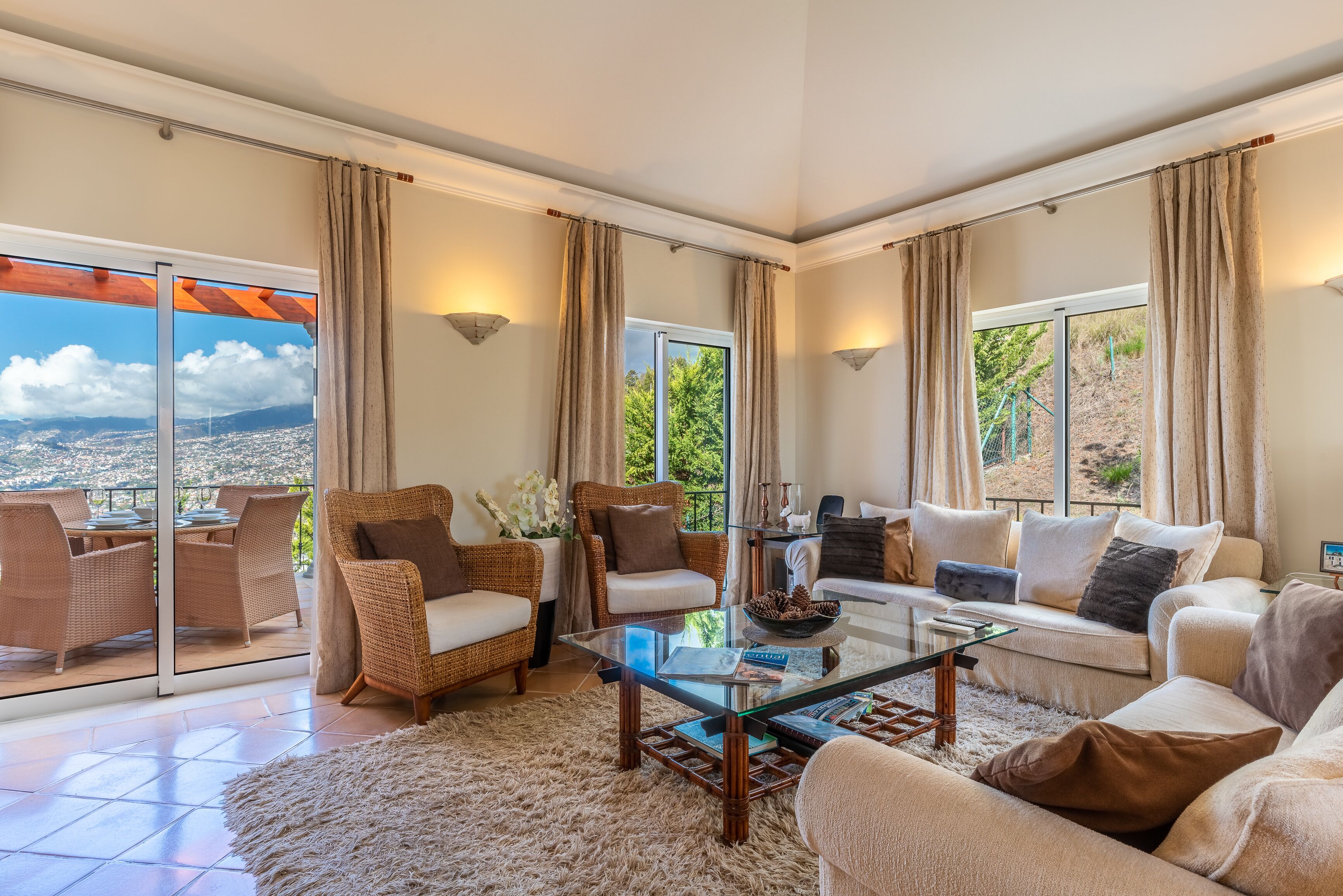 A spacious living room with a glass coffee table, wicker furniture, and large windows offering a city view.
