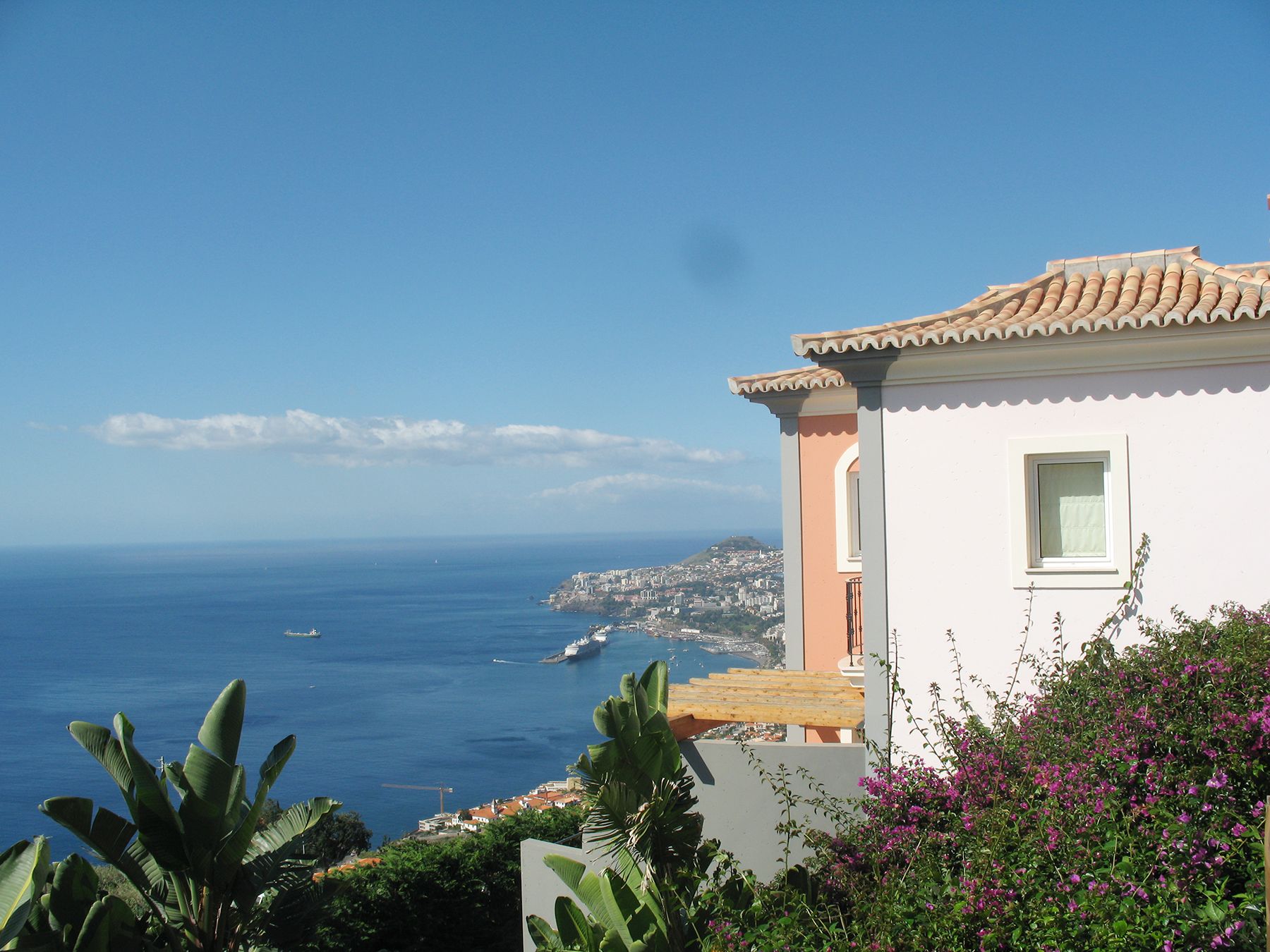 A coastal building with a white facade and terracotta roof tiles, overlooking a clear blue sea and a distant cityscape.