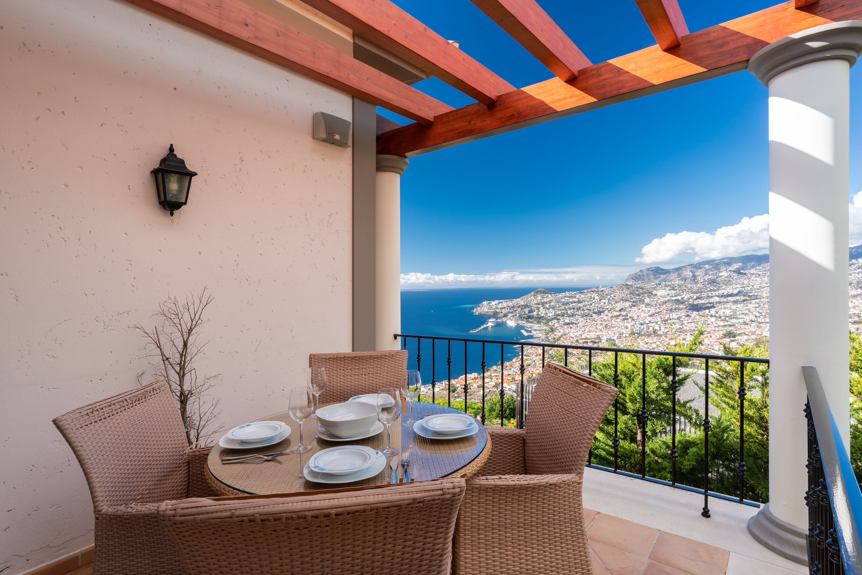 A balcony with wicker furniture, a table set for dining, and a view of a coastal city.
