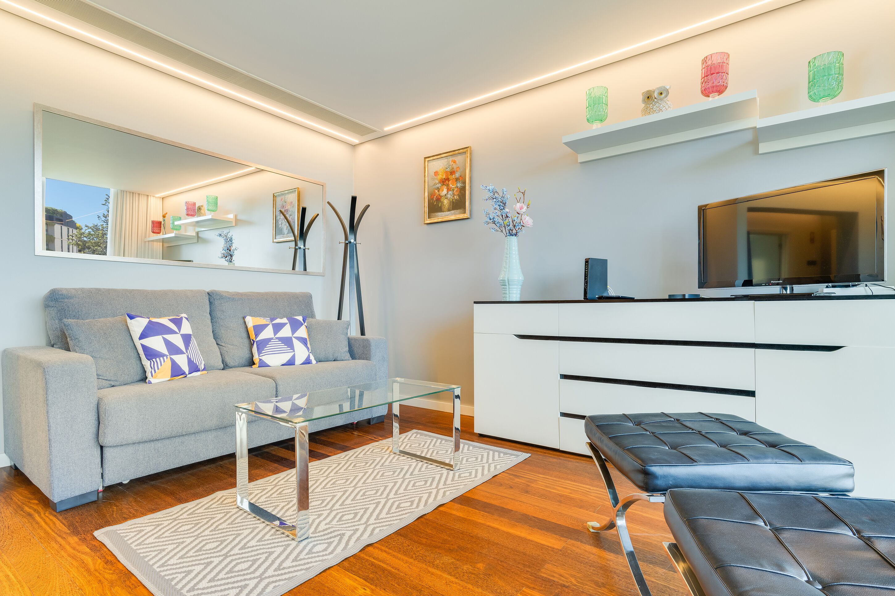 A modern living room with a grey sofa, a glass coffee table, a black ottoman, a white cabinet, and a large flat-screen TV.