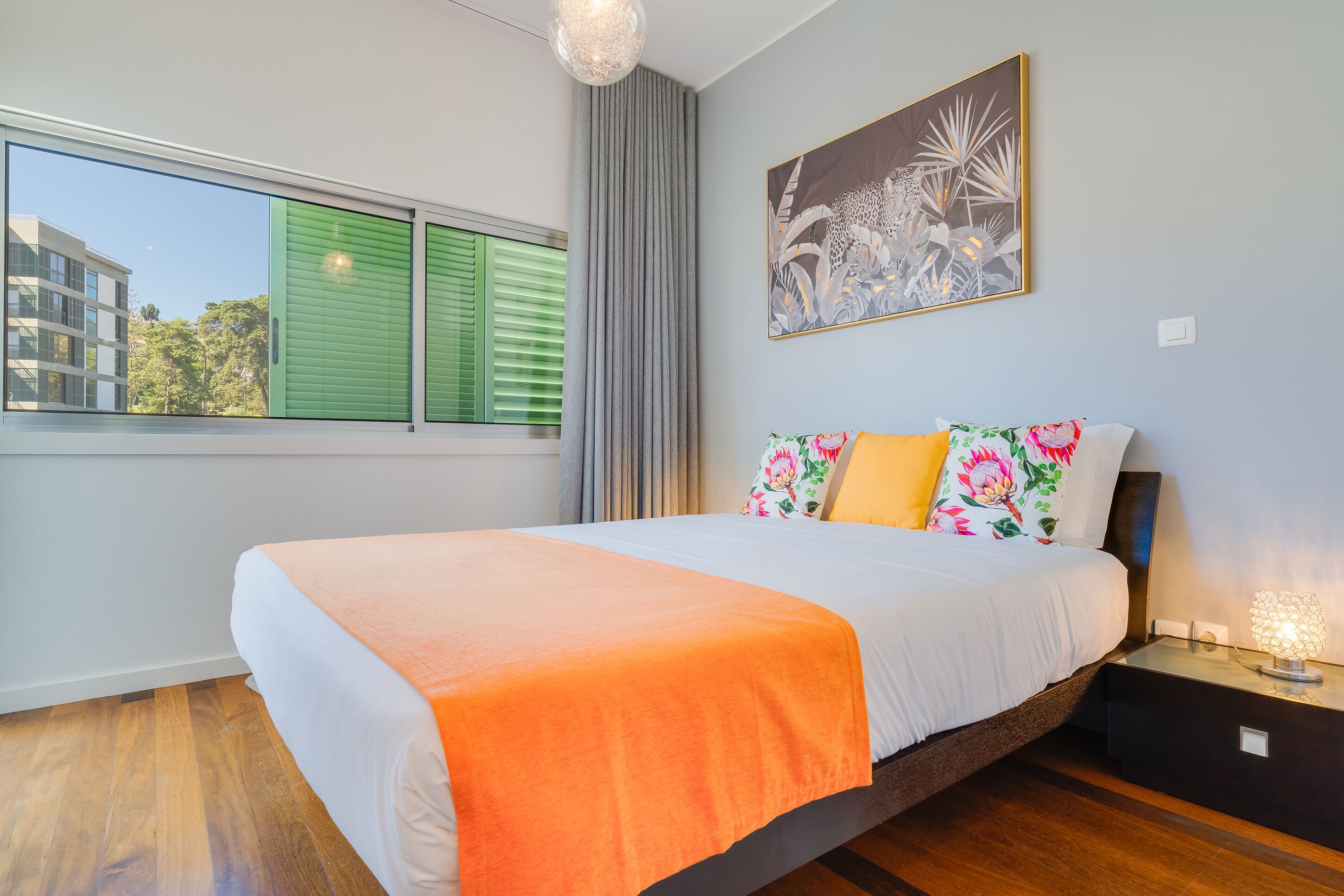 A modern bedroom with a large bed, a window with green shutters, a city view, and a floral artwork on the wall.
