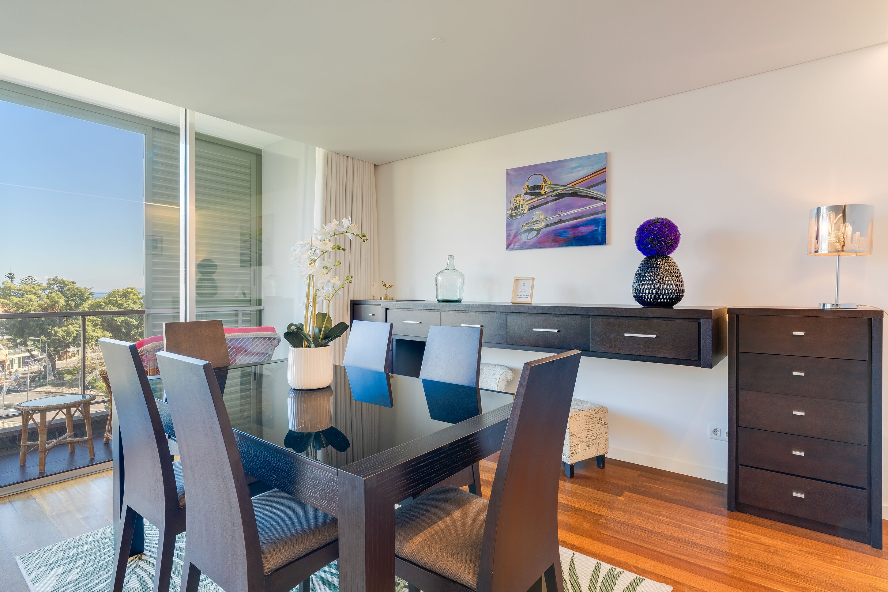 A modern dining area with a glass table, dark chairs, and a view of the outdoors.