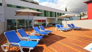 A rooftop deck with blue lounge chairs and umbrellas, a wooden deck, and a modern building with green shutters. - Villa Nogueira IV (Funchal)
