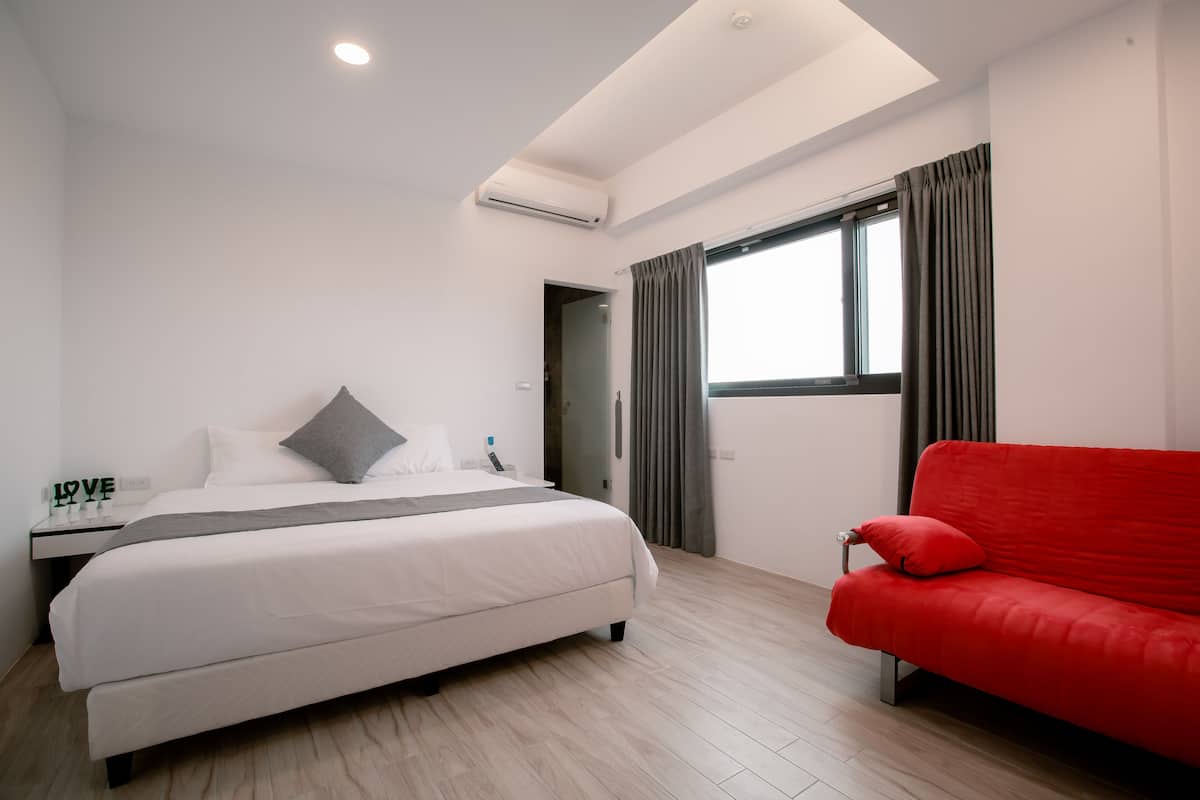 A modern hotel room with a bed, a red sofa, a window with curtains, and a skylight.