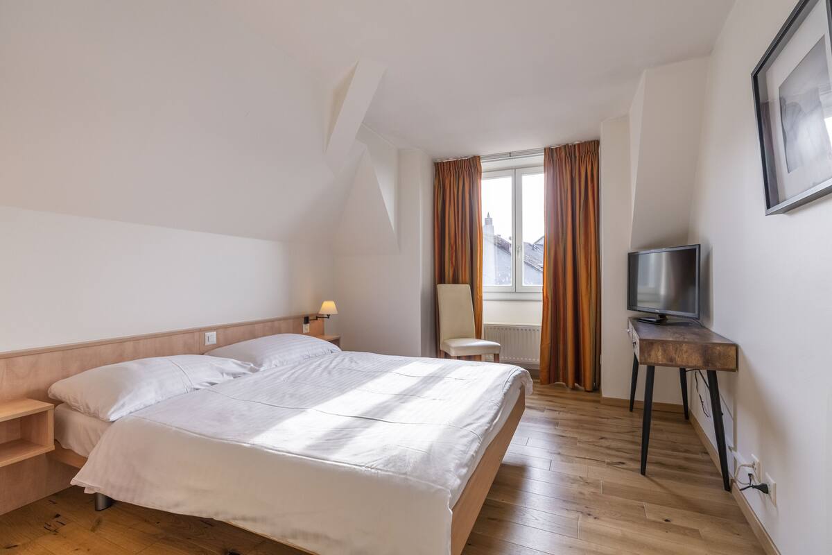 Quarto de hotel com cama, televisão sobre suporte de madeira, uma cadeira e janela com cortinas.