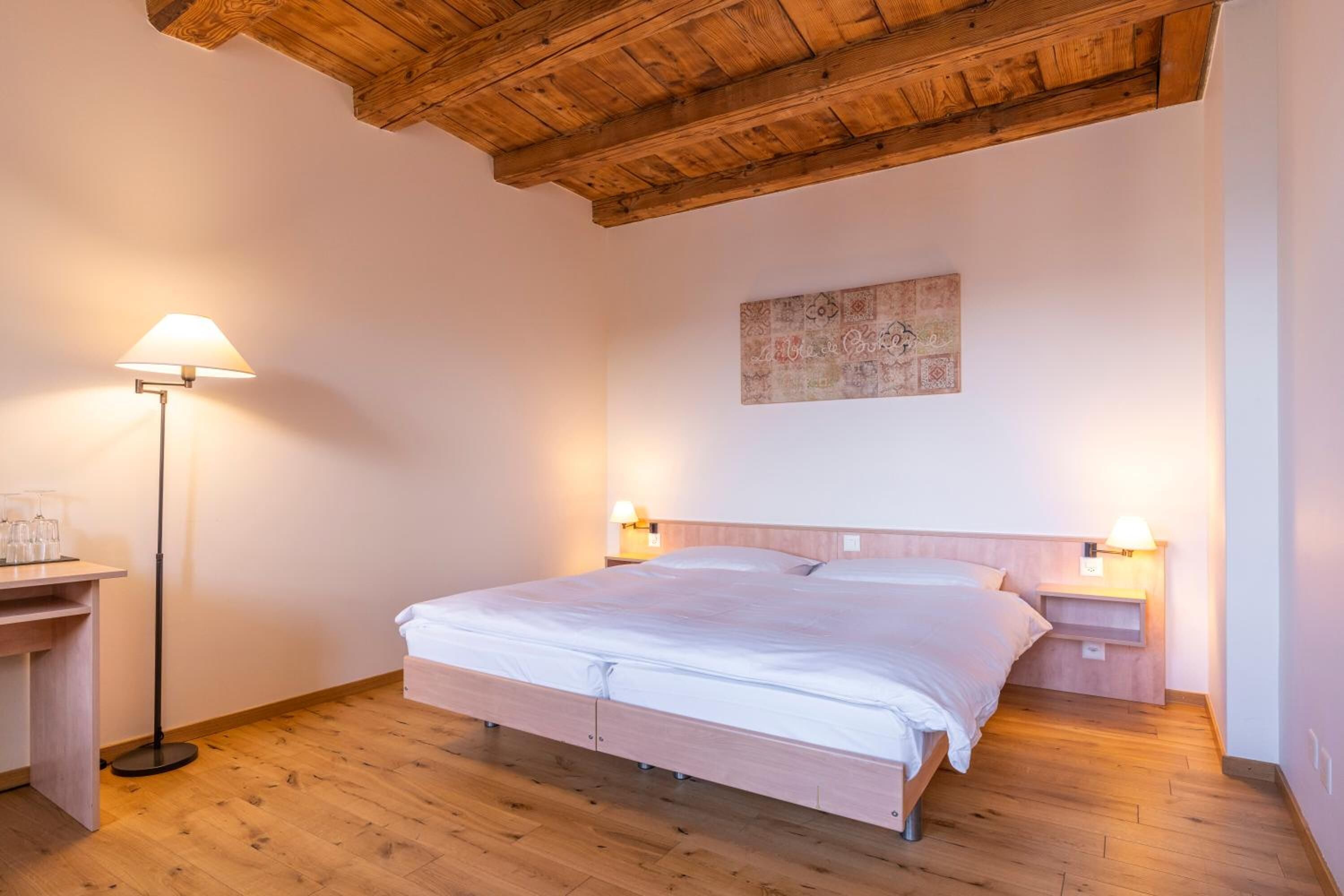 Een slaapkamer met een houten plafond, een bed met wit beddengoed, twee nachtkastjes met lampen, en een klein bureau met een lamp.