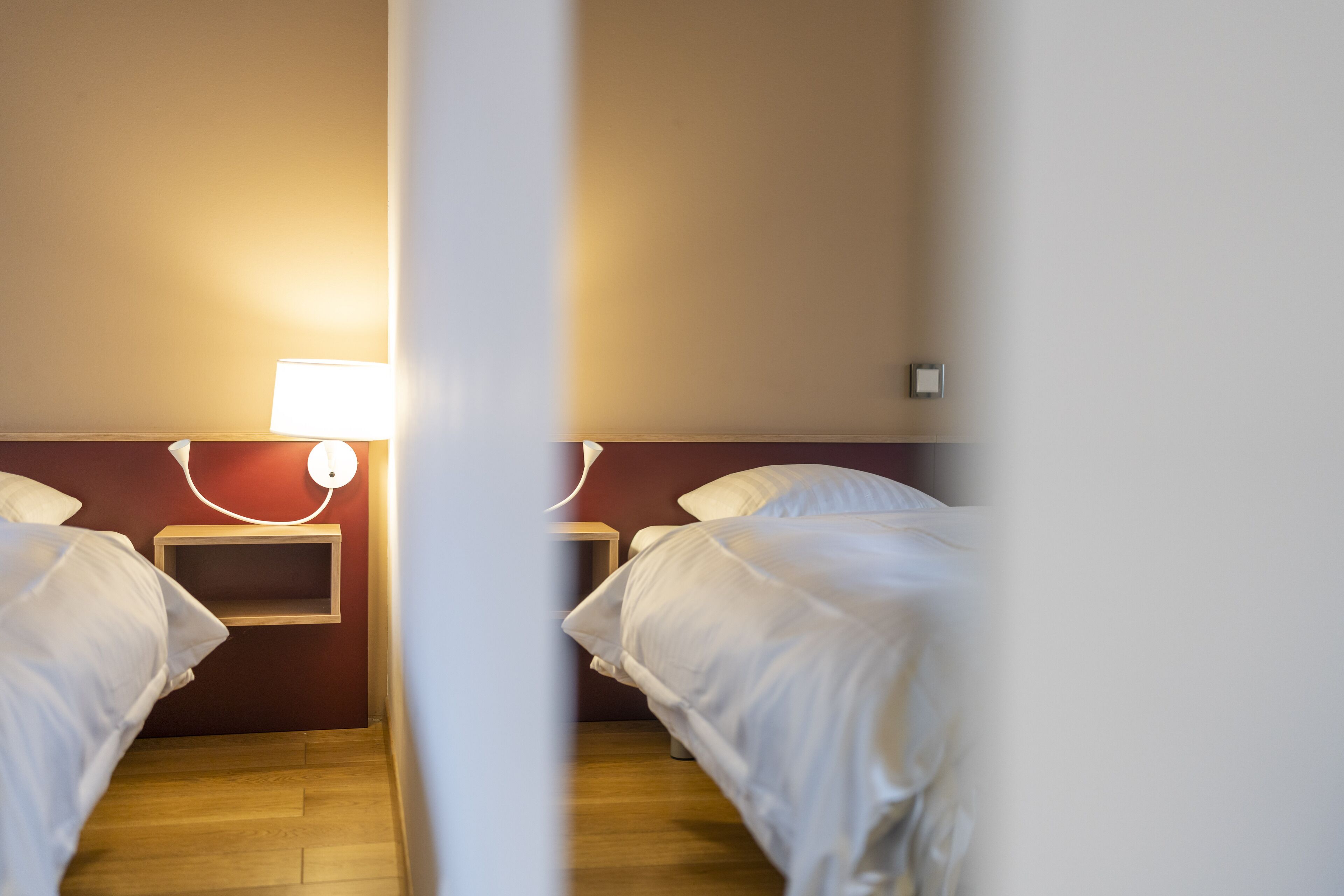 A hotel room with two beds, a bedside table, a nightstand, and a wall-mounted lamp.