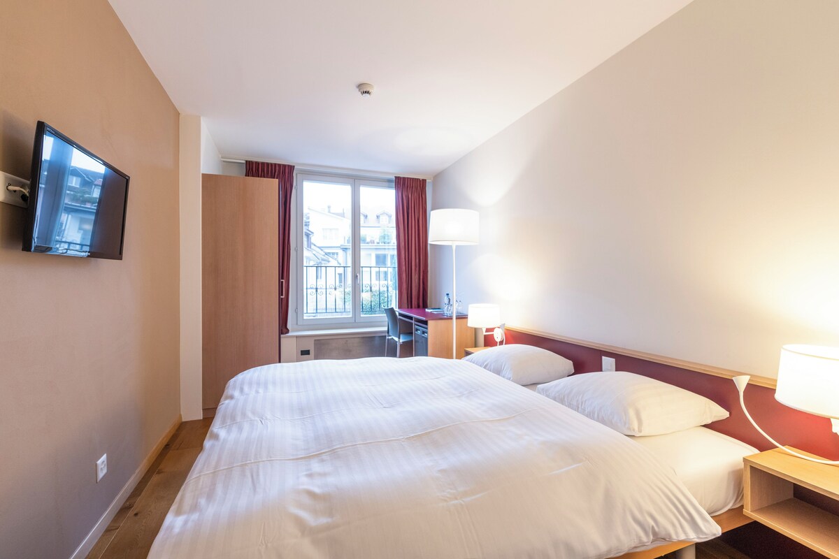 A hotel room with a bed, a desk, a chair, a TV mounted on the wall, and a window with curtains.