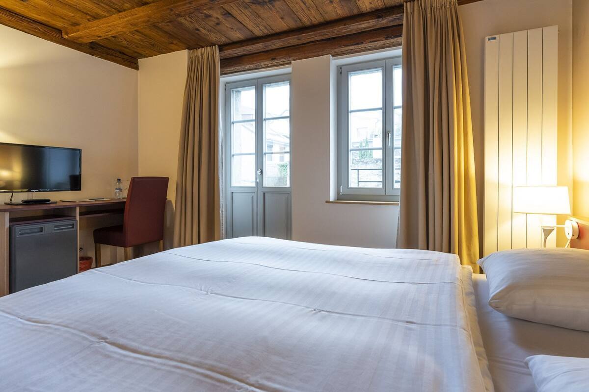 Quarto de hotel com cama, escrivaninha com cadeira, televisão e janela com cortinas.