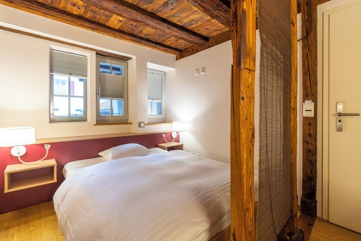 A bedroom with a wooden ceiling, two bedside tables with lamps, a bed with white bedding, and a window with blinds.
