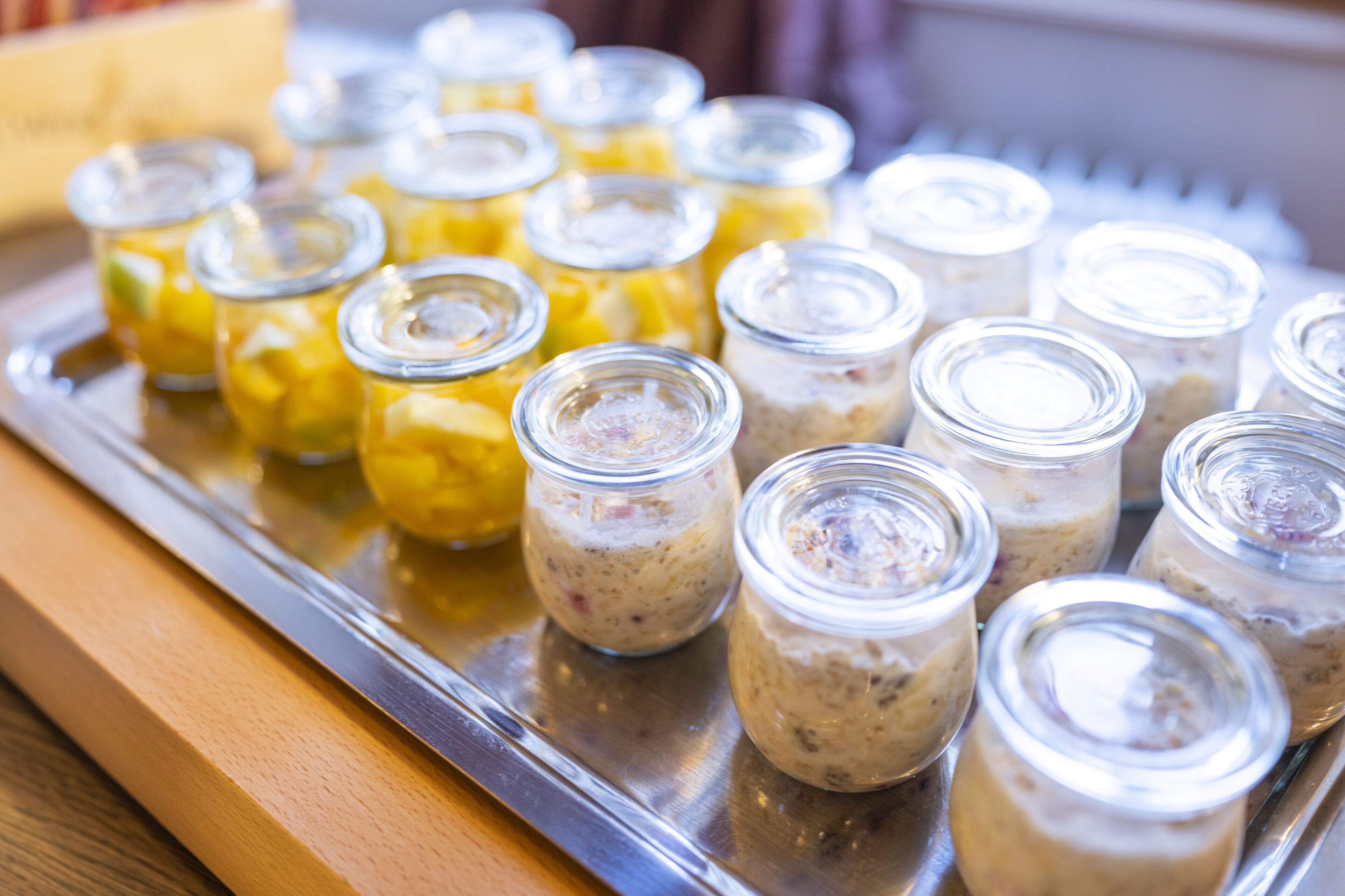 Un plateau contenant divers aliments, notamment des bocaux avec des contenus jaunes et bruns, qui pourraient être des cornichons ou des confitures, le tout posé sur un plateau en métal.