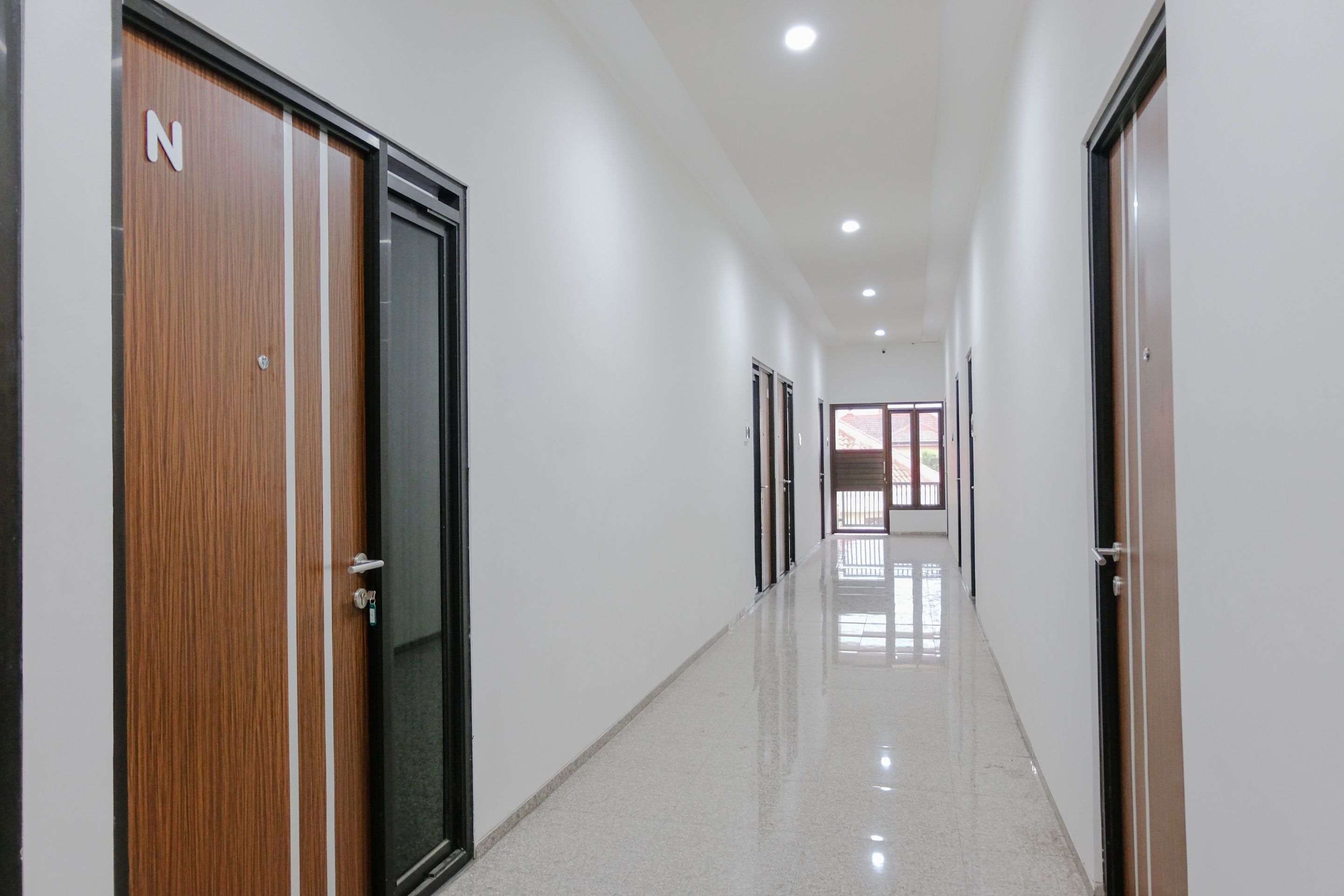 Um corredor comprido e estreito, com portas de madeira de um lado e paredes brancas.