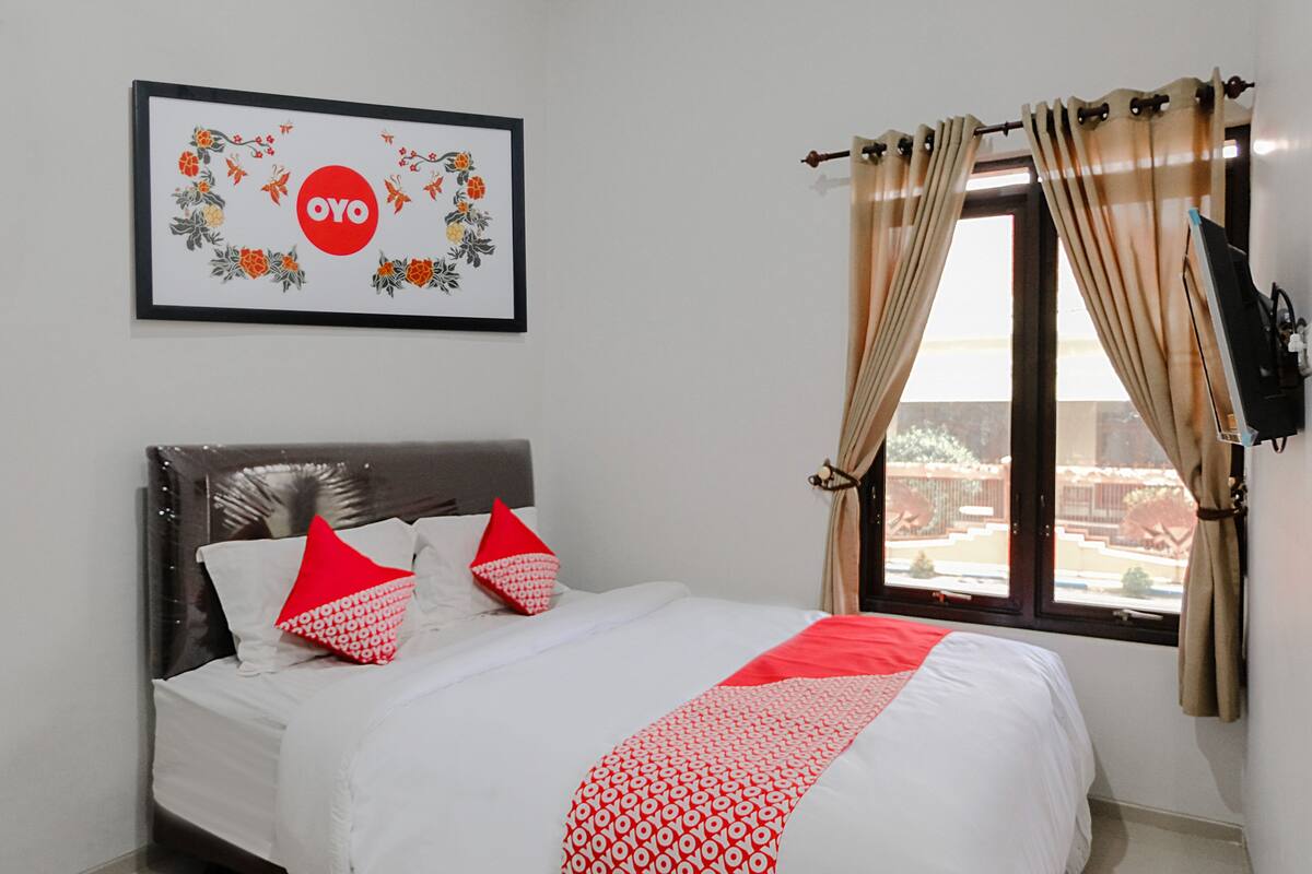A neatly made bed with white and red bedding, a framed artwork with floral and text design, and a window with curtains.