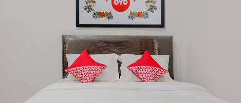Cama bem arrumada com roupa de cama branca e vermelha, duas almofadas com padrão repetido e um quadro com logotipo e design floral na parede acima.