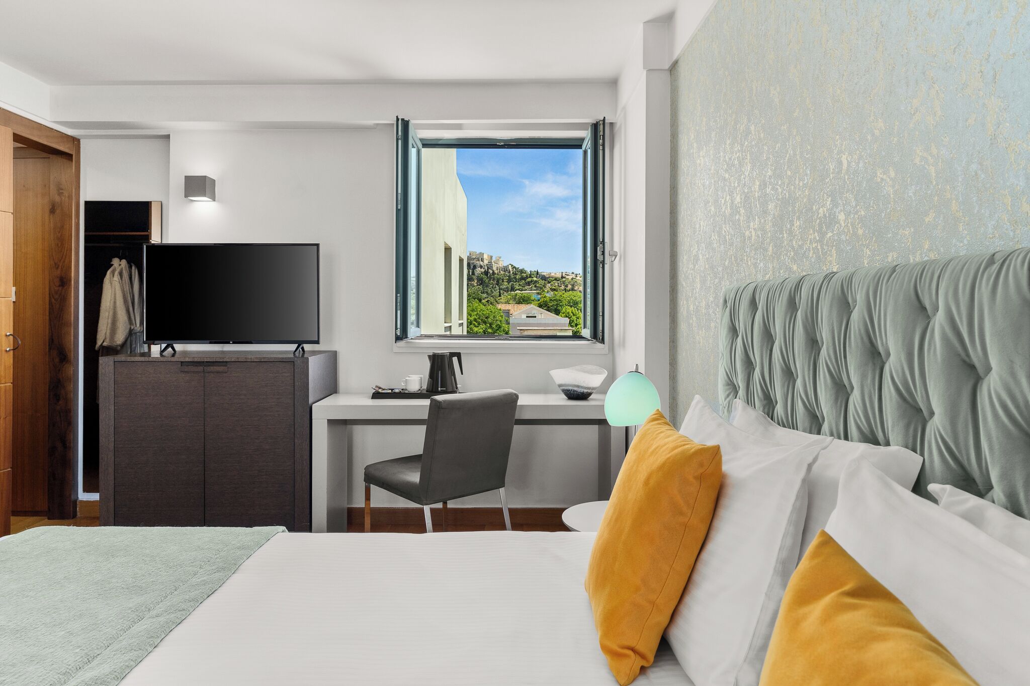 A hotel room with a bed, a desk with a chair, a television, and a window with a view of greenery and buildings.