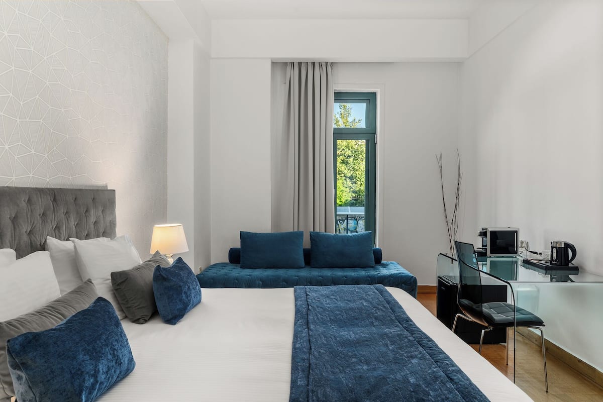 A modern hotel room with a large bed, a desk, and a view of greenery outside.