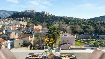A table set with plates of food, glasses, and a vase of yellow flowers on a balcony overlooking a town with historic buildings and a hill in the background.