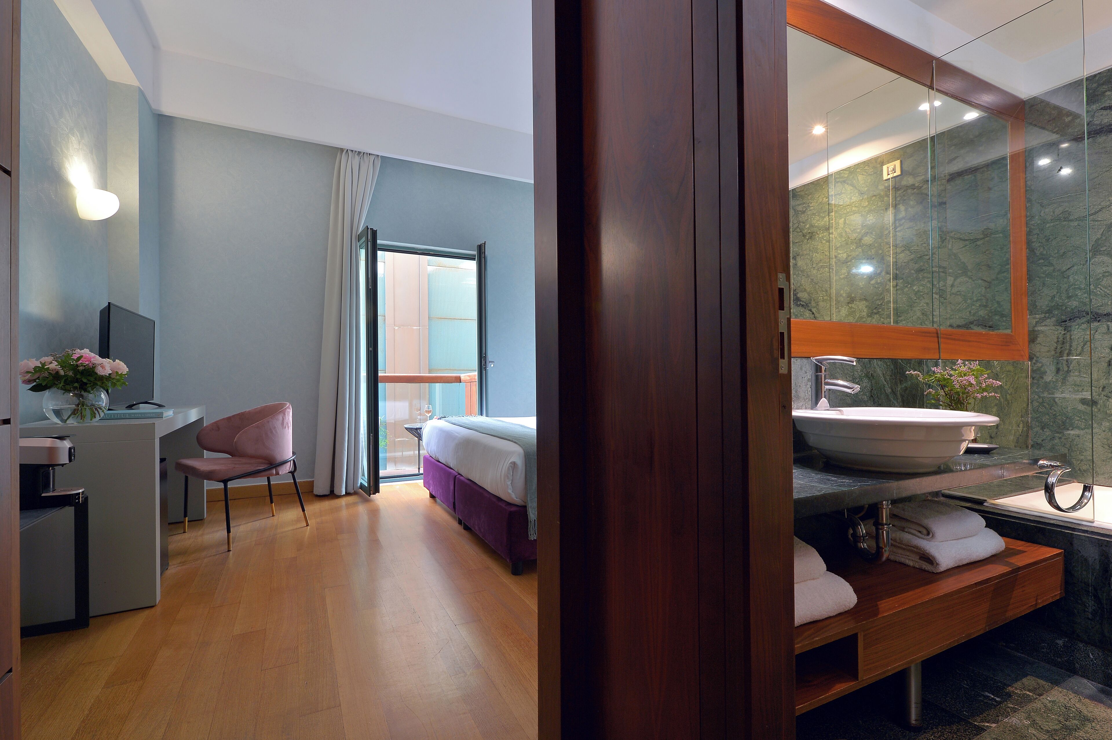 A modern hotel room with a wooden vanity, a large mirror, a bed, a desk, and a chair.