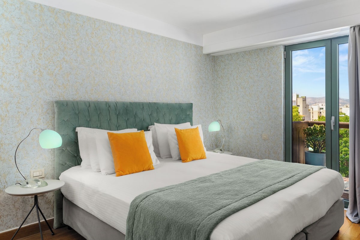 A hotel room with a bed, a nightstand, a lamp, and a view of the city through a large window.