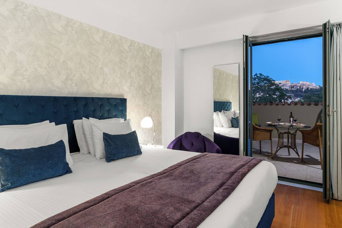 A hotel room with a large bed, a view of a balcony, and a table with chairs outside.