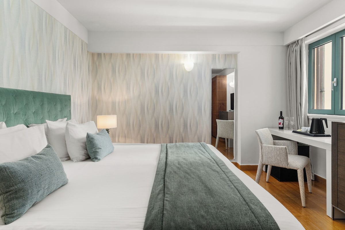 A hotel room with a bed, a desk, a chair, a lamp, and a window.