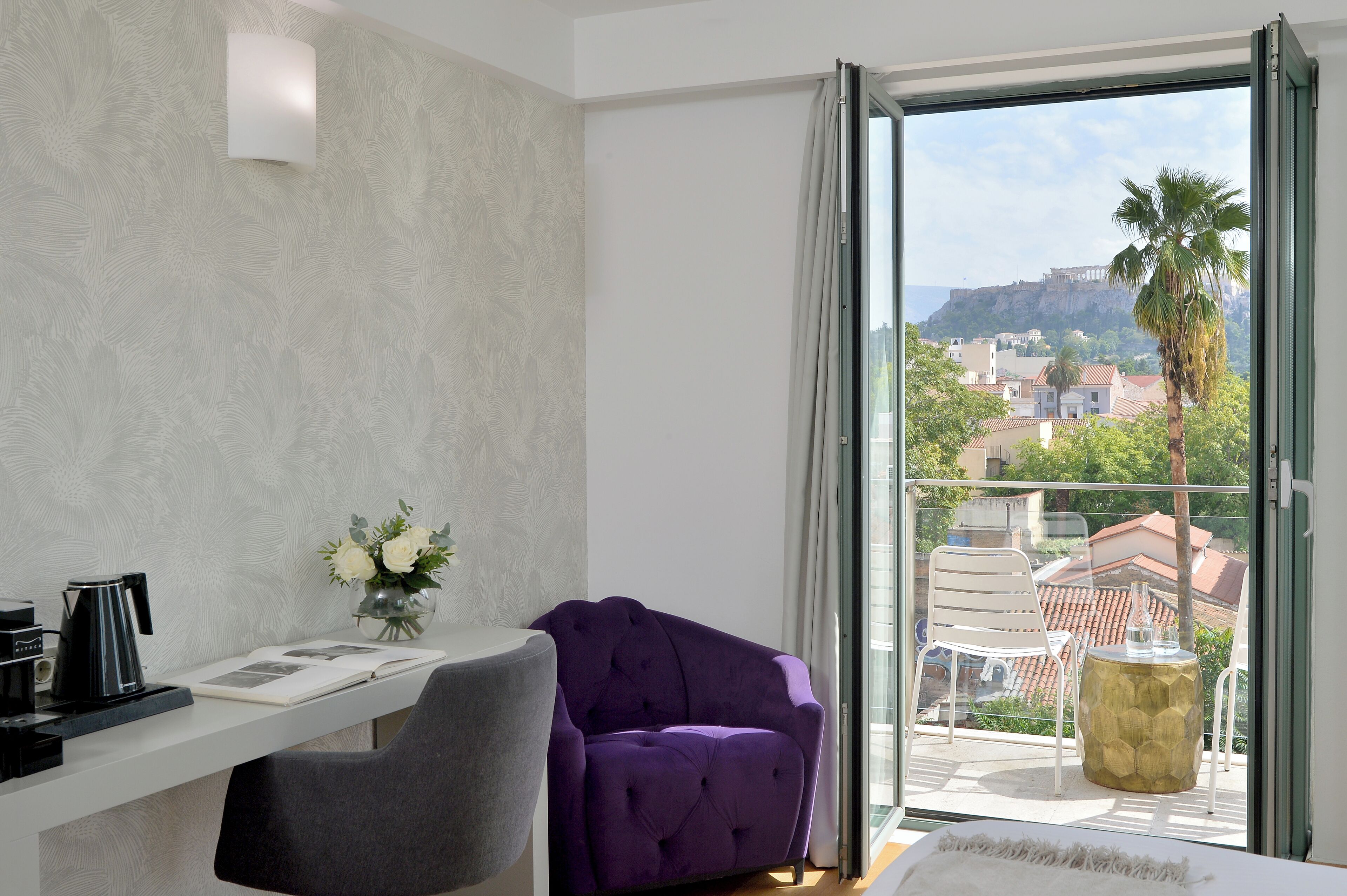 Une chambre moderne avec un bureau, une chaise violette, une chaise blanche, une petite table ornée d’un vase de fleurs et une vue sur le paysage urbain, visible à travers une porte vitrée.